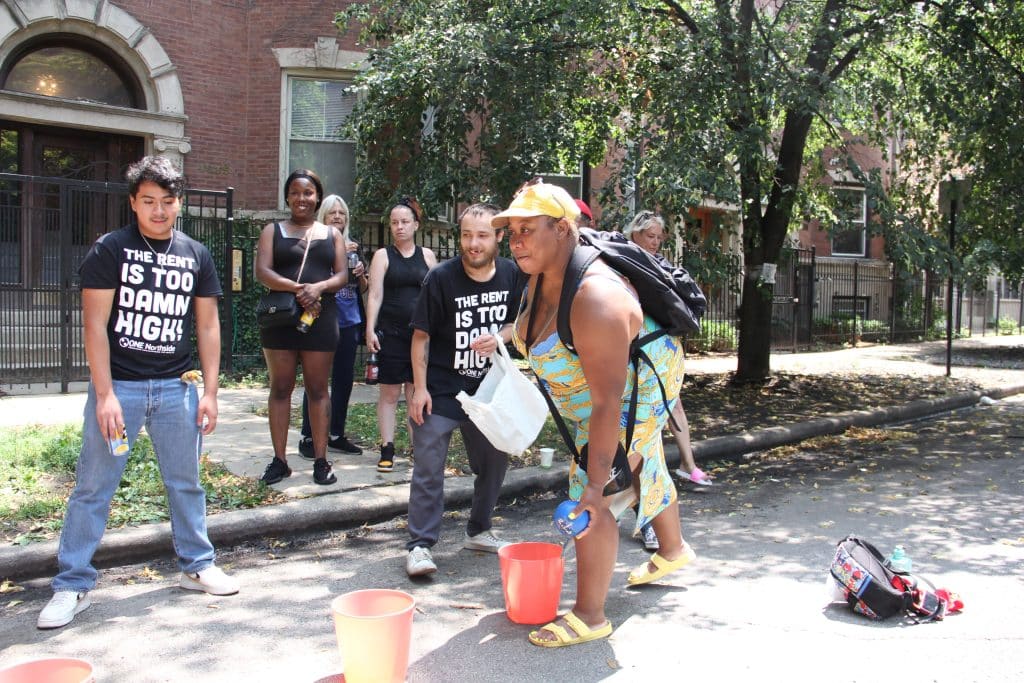 Woman in Summer dress throwing a ball into buckets, as several people watch. Two of the people watching are wearing black t-shirts that read "The Rent is Too Damn High"