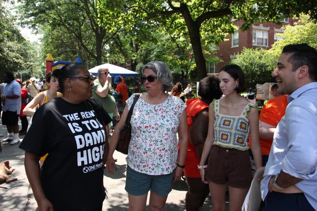 In the foreground, 4 people talk. One is wearing a black t-shirt that says "The Rent is Too Damn High." In the background are many people walking around and having fun, under a canopy of trees.
