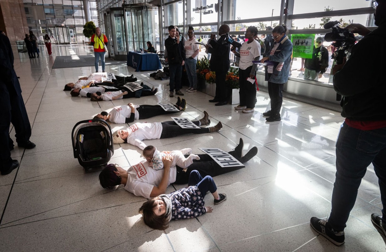 7 Adults (and two children) lay on the ground in a Die-In, each individual adult representing 100,000 per day claims who are denied by private insurance companies. The people on the ground are surrounded by folks taking pictures as well as activists speaking