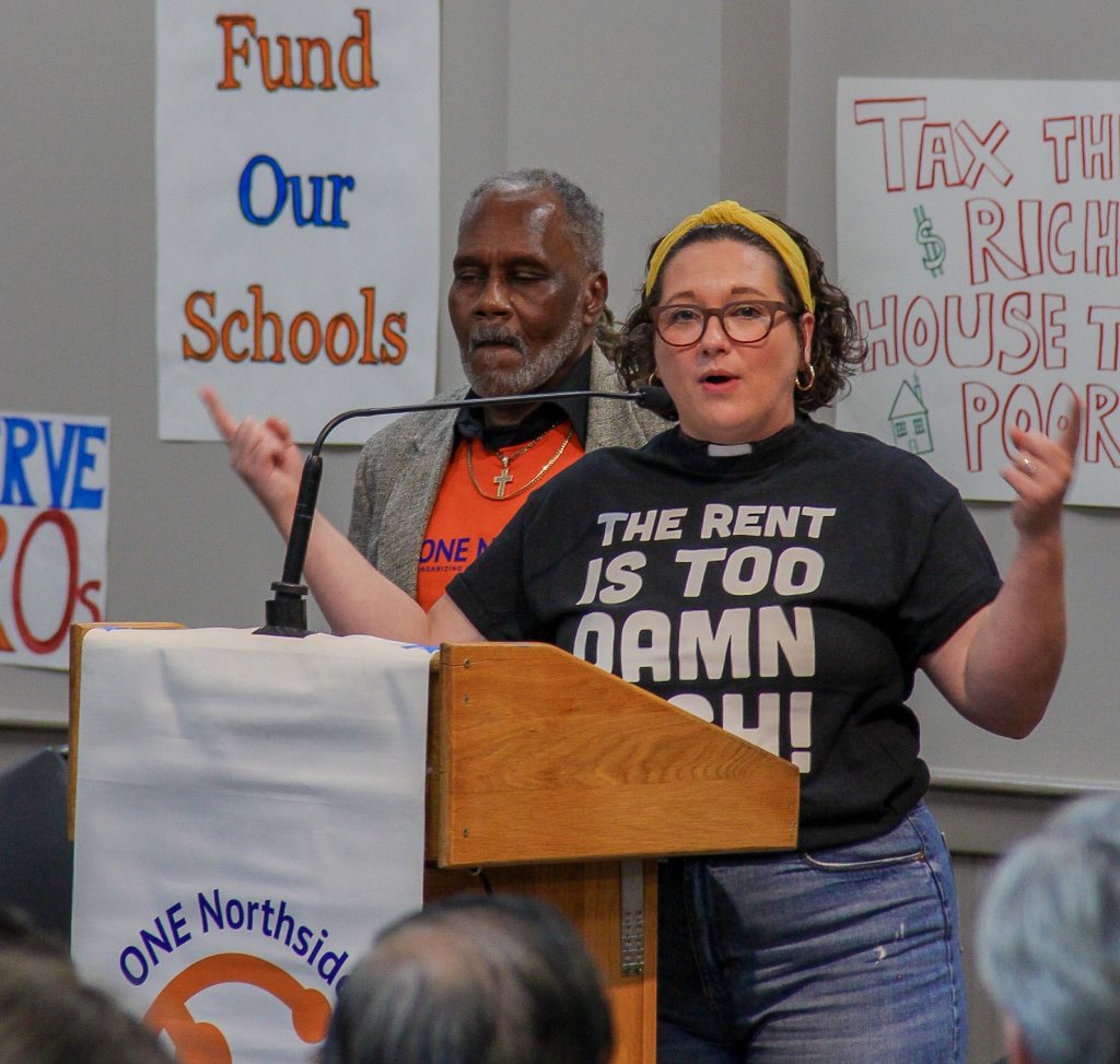 Lindsey at the podium with Anthony just behind her, protest signs on the wall behind them.