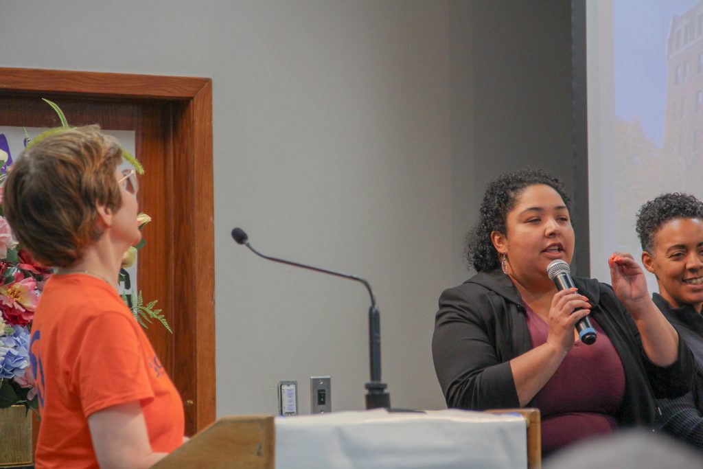 Sue at the podium turns to watch Commissioner Castaneda speaking into a microphone addressing the audience