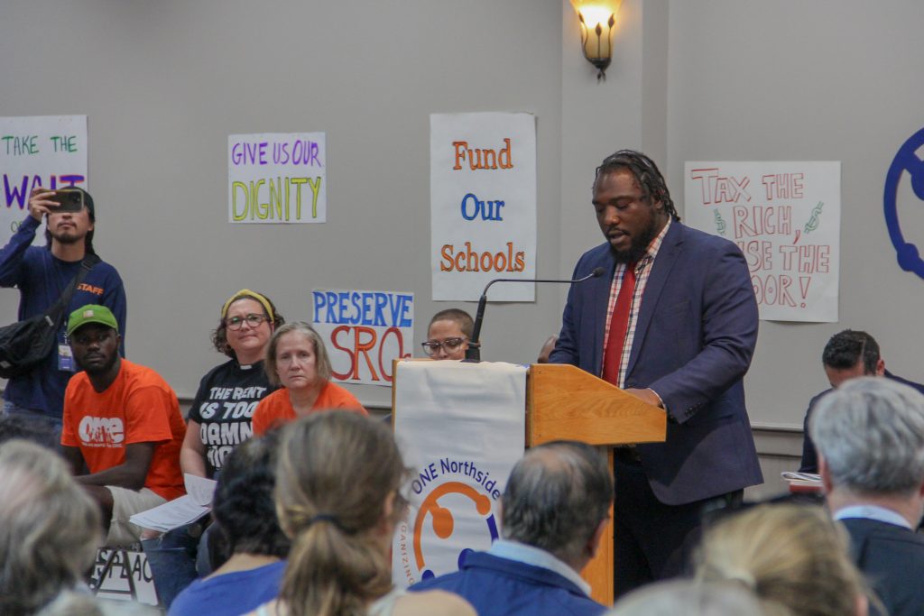 Darrell speaks at the podium while others wait behind him and some of the crowd can be seen in the foreground