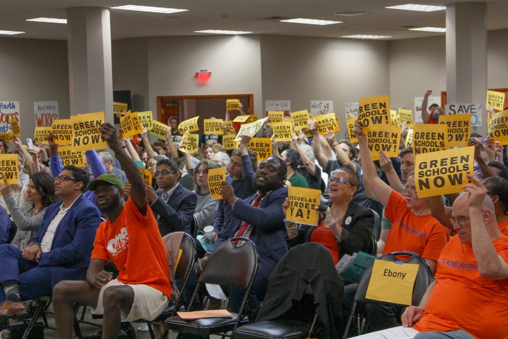 An audience holding signs reading Green Schools Now