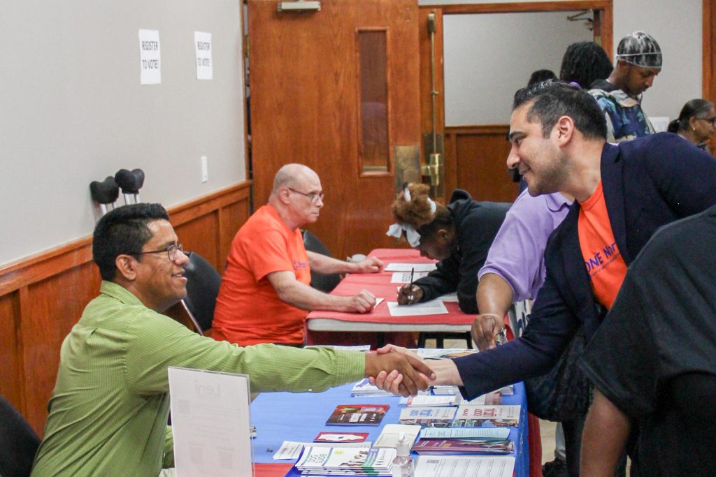 Jesse leaning over a table to shake hands with Brock, Kerry at a table in the background